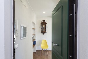 Un pasillo estrecho con una puerta verde, un reloj de madera, una silla amarilla y un teléfono.