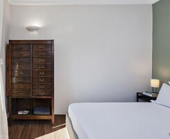 Un dormitorio con una cama, un aparador de madera con varios cajones, una mesita de noche con una lámpara y un accesorio de luz instalado en la pared.