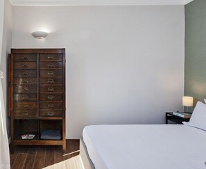 Une chambre d’hôtel comprenant un lit, une commode en bois avec plusieurs tiroirs, une table de chevet avec une lampe et un luminaire fixé au mur.