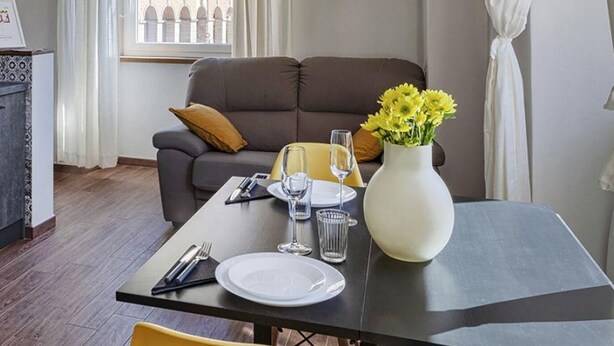 A modern living room with a sofa, a dining table set for two, and a vase of yellow flowers. - San Lorenzo View Apartment 5 by Wonderful Italy (Genoa)