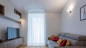 A modern living room with a grey sofa, a flat-screen TV, a wooden cabinet, and a large window with sheer curtains.