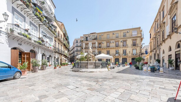 A paved square with a central fountain, surrounded by multi-story buildings with balconies and potted plants.
