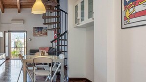 A spacious room with a wooden herringbone floor, a dining area with a glass table and chairs, a staircase with a wooden railing, and a large framed artwork on the wall.
