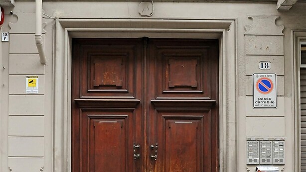 A large, wooden double door with a metal handle, set in a building with a light-colored facade and a small balcony above.