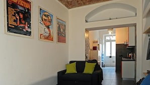 A living room with a black sofa and green cushions, a kitchen with a white fridge, and a doorway leading to another room.