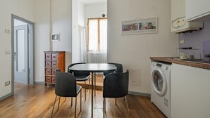 A compact kitchen with a washing machine, a dining table with chairs, and a cabinet with decorative boxes.