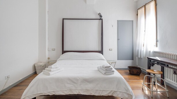 A minimalist hotel room with a large bed, a wooden floor, a radiator, and a window with curtains.