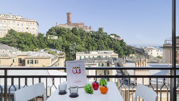 Un balcón con una mesa que incluye un cartel de bienvenida, un vaso y frutas decorativas. Se aprecia un edificio histórico con una torre al fondo.