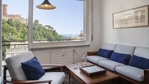 Una sala de estar con un sofá, un sillón y una mesa de centro, un ventanal con vistas a una colina y edificios, y un cuadro en la pared.