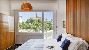 Un dormitorio con un ventanal grande, una cama con ropa de cama blanca y azul, un armario de madera y vistas a un castillo.