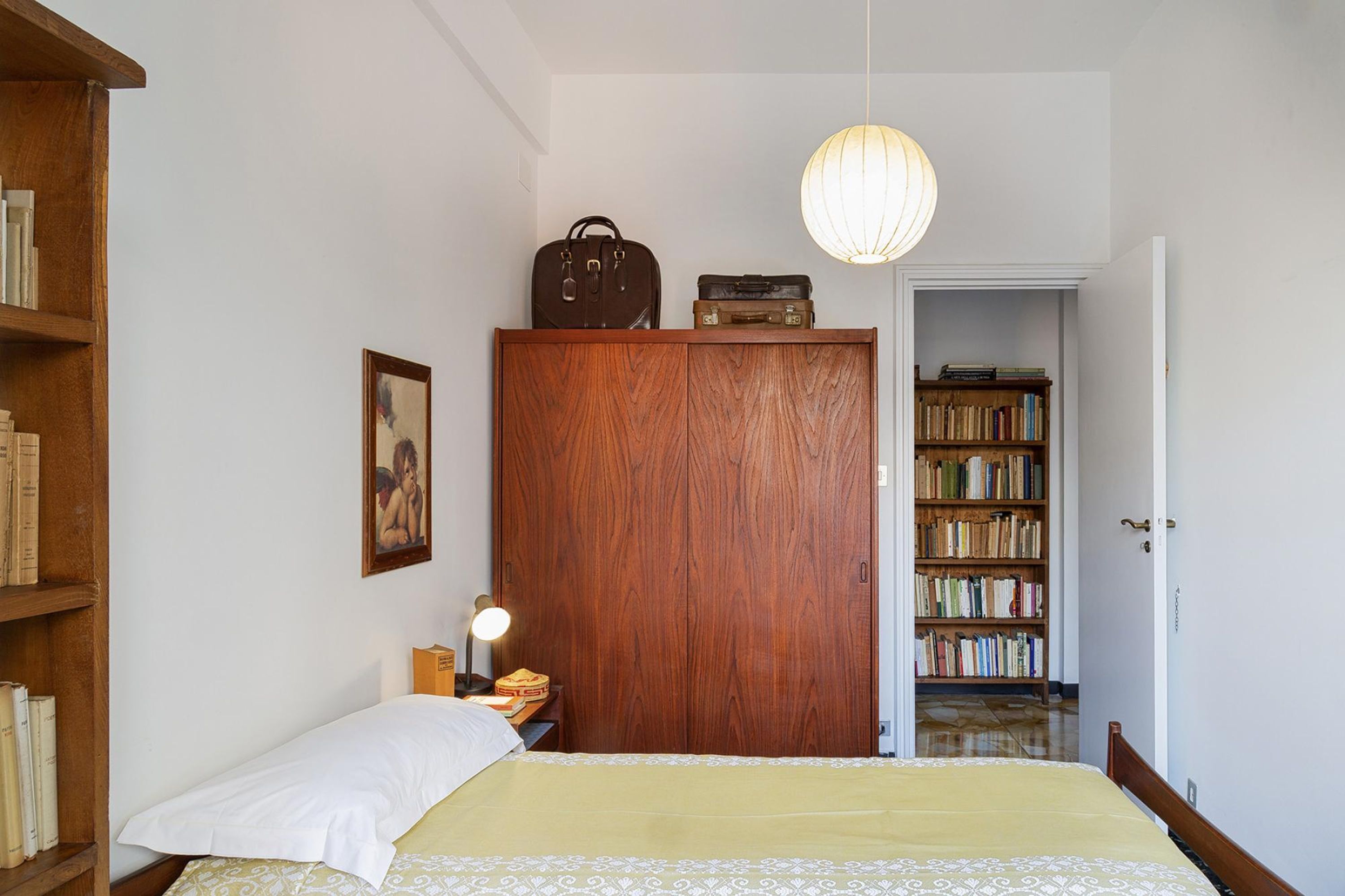 A bedroom with a bed, a wooden wardrobe, a bedside table with a lamp, a hanging light fixture, and a bookshelf filled with books.