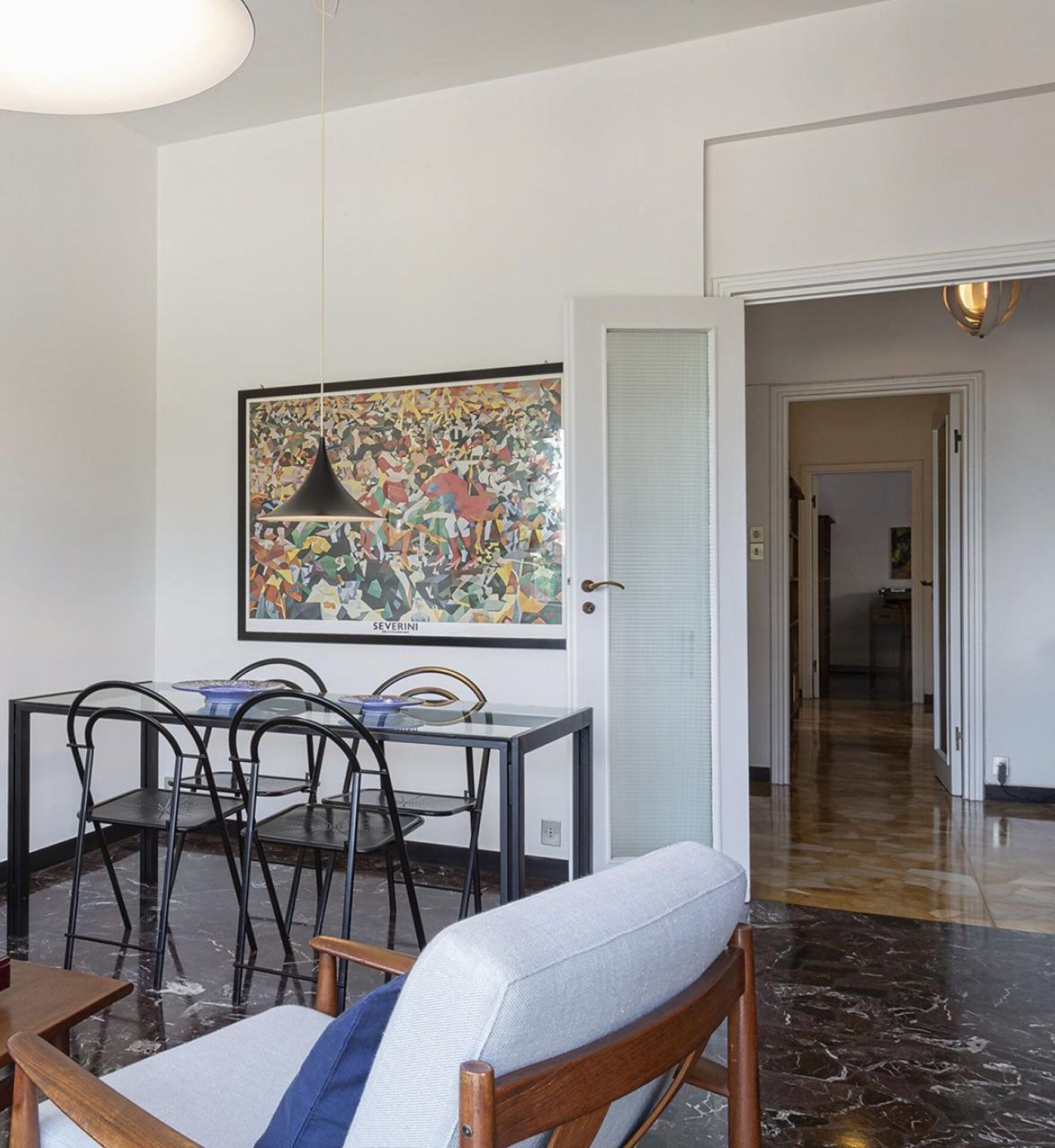 Un comedor con una mesa y sillas de vidrio, un cuadro abstracto enmarco y un pasillo que conduce a otra habitación.