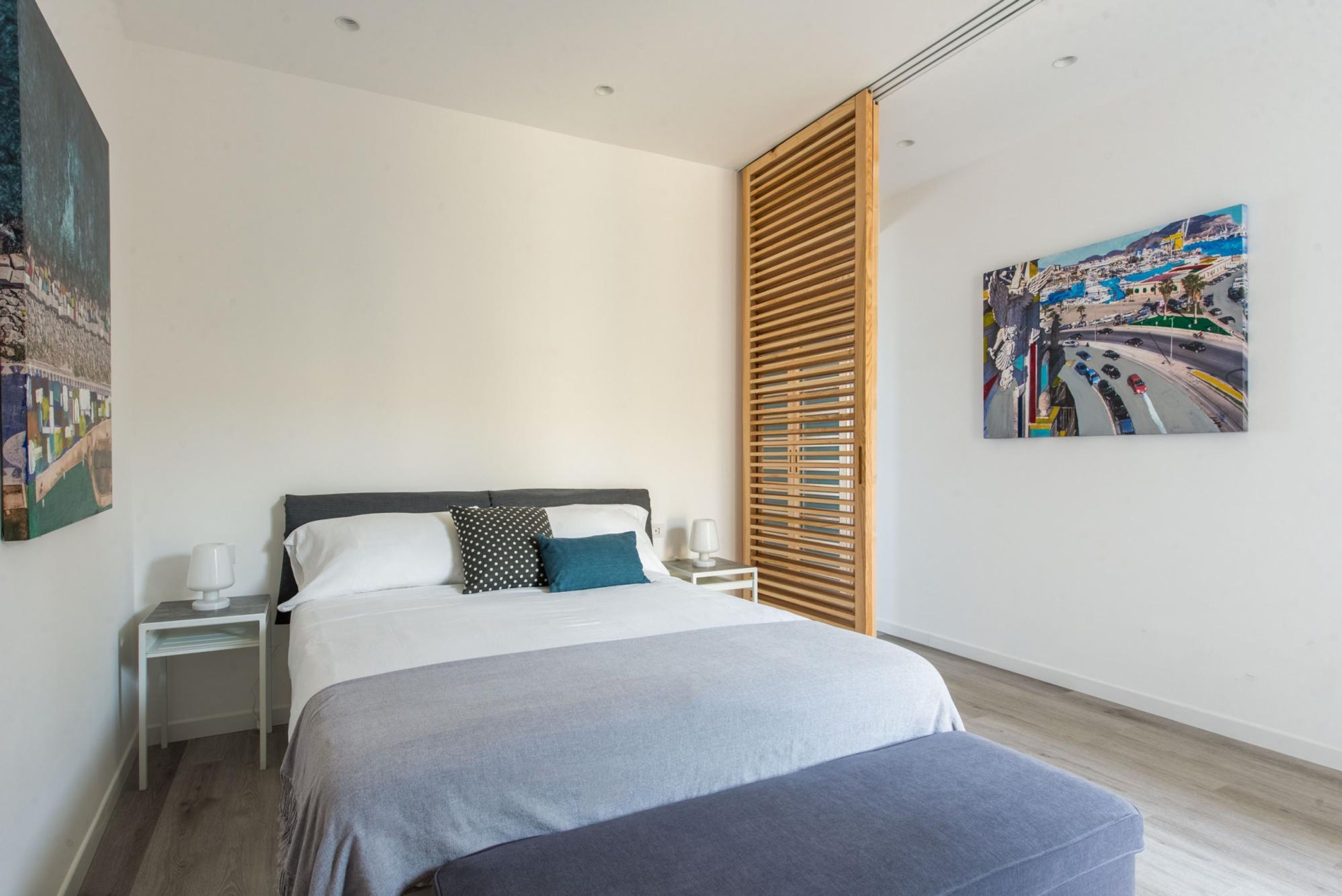 A modern bedroom with a large bed, a nightstand, a wooden slat partition, and a cityscape painting on the wall.