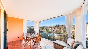 A balcony with a dining table and chairs, a sofa, and a view of a swimming pool and houses. - La Piopa 2 20 in Polpenazze del Garda (Polpenazze del Garda)