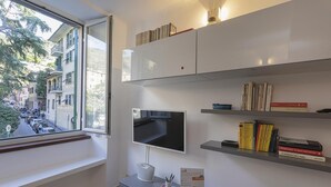 A modern room with a large window, a flat-screen TV, a desk with a basket, and shelves with books.