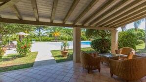 A covered patio area with wicker furniture, a pool, and lush greenery in the background. - Villa Arenella con Piscina by Wonderful Italy (Syracuse)