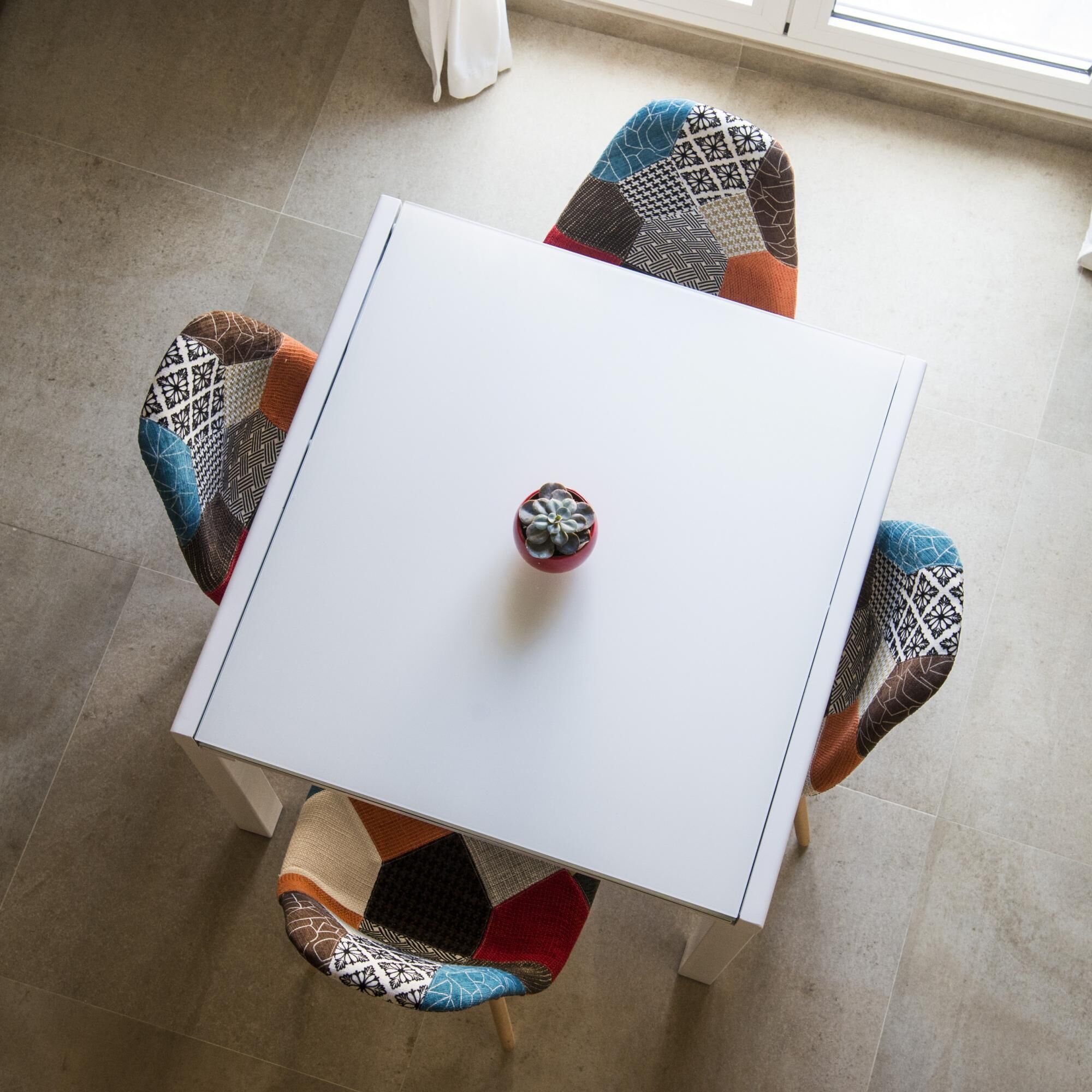 A white square table with a red decorative item in the center, surrounded by four chairs with patterned upholstery.