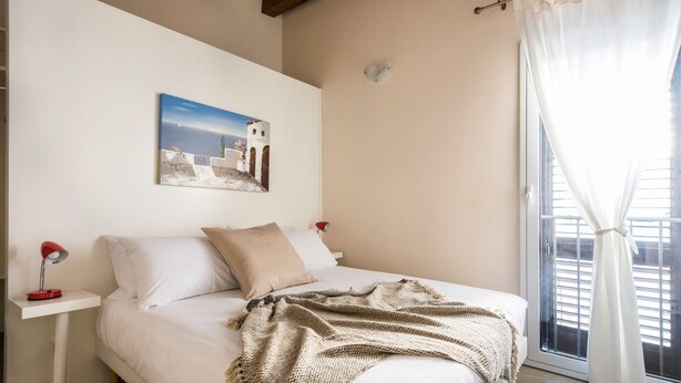 A bedroom with a bed, a nightstand, a lamp, a painting, and a window with curtains.