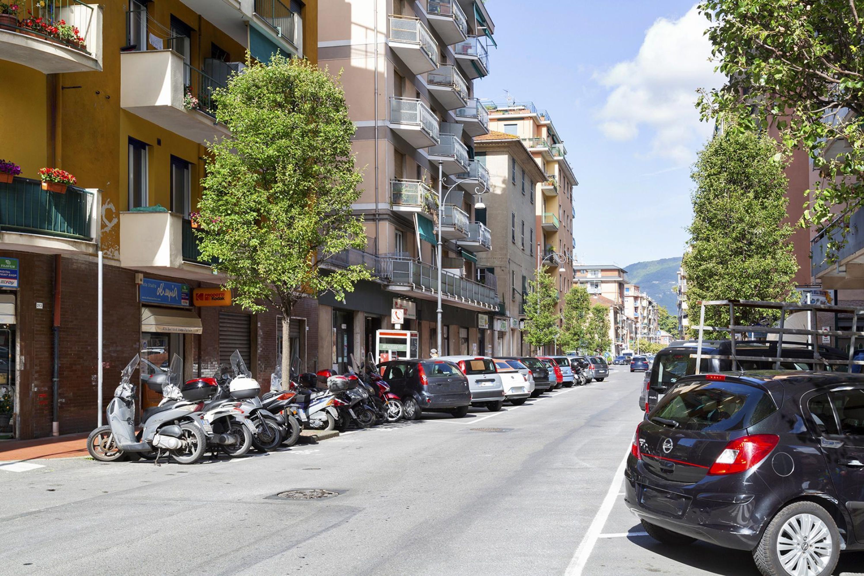 Eine Straße mit geparkten Autos und Motorrädern, mehrstöckige Gebäude mit Balkonen und einem wolkenlosen Himmel.