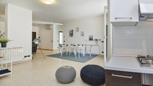 Une cuisine moderne avec des armoires blanches et une hotte de cuisinière en acier inoxydable. On y trouve un coin repas avec une table et des chaises blanches, ainsi qu’un petit tapis sur le sol.