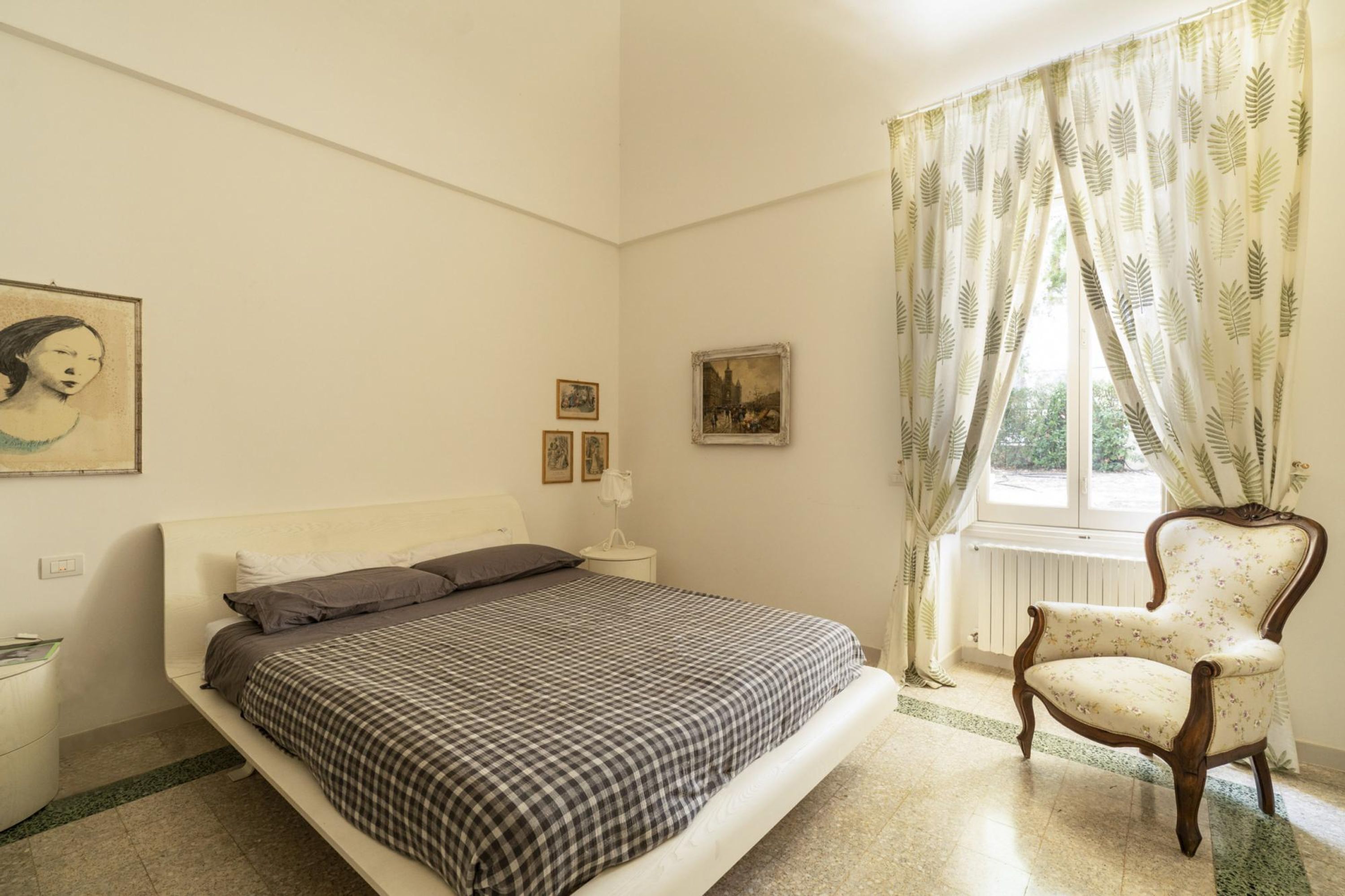 Una camera da letto con un letto, una sedia, una finestra con tende e quadri appesi al muro.