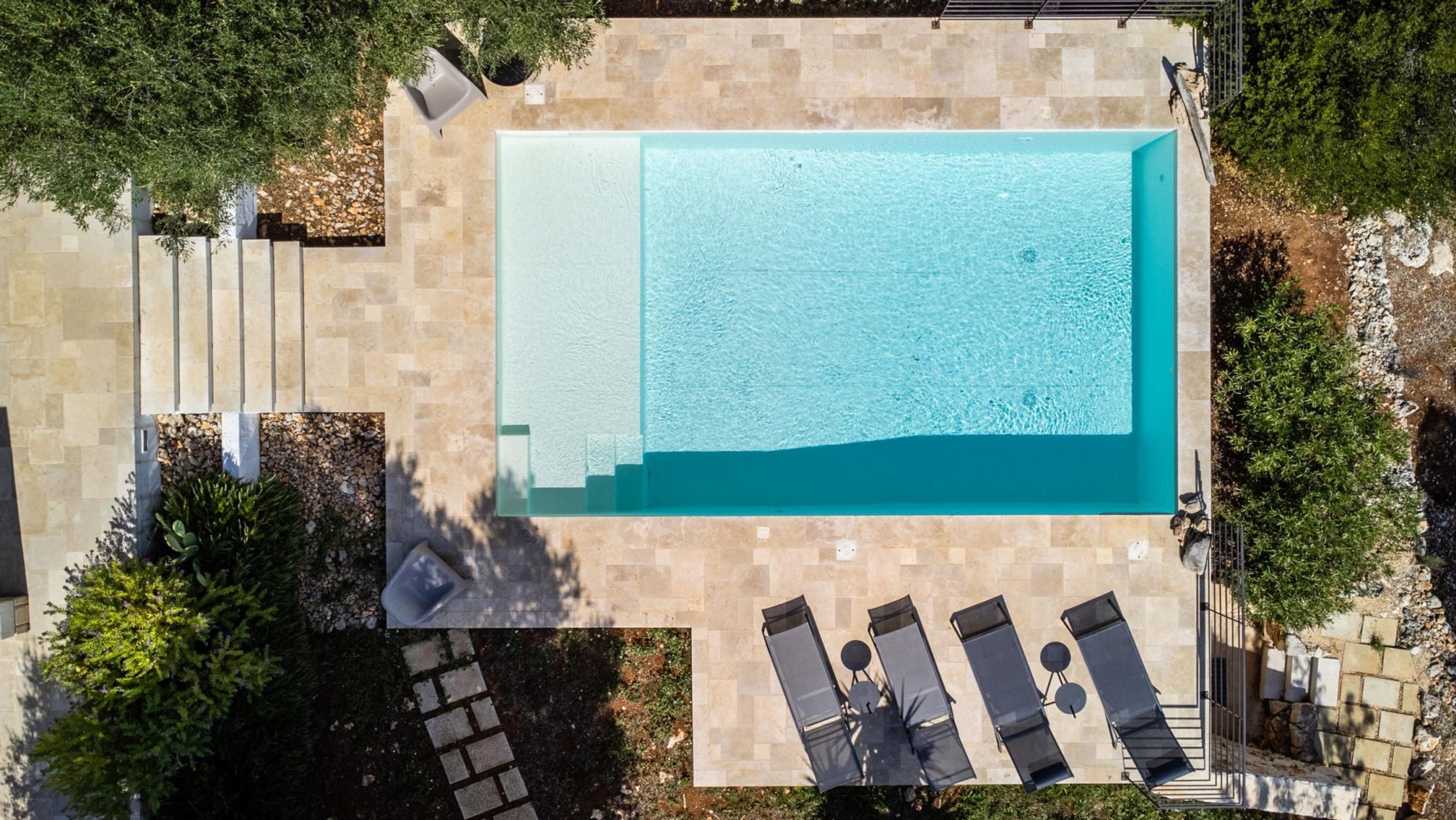 An aerial view of a rectangular swimming pool surrounded by stone tiles, with lounge chairs and a small table nearby.
