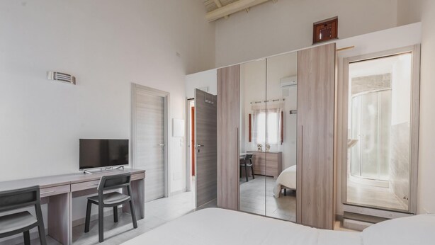 A modern hotel room with a bed, a desk with a chair, a TV, and a bathroom visible through a glass door.