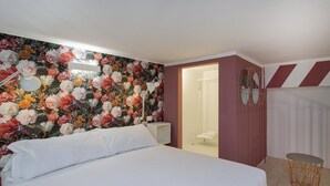 Habitación de hotel con papel tapiz floral, una cama con sábanas blancas, una mesita de noche pequeña, un espejo y una pared a rayas.