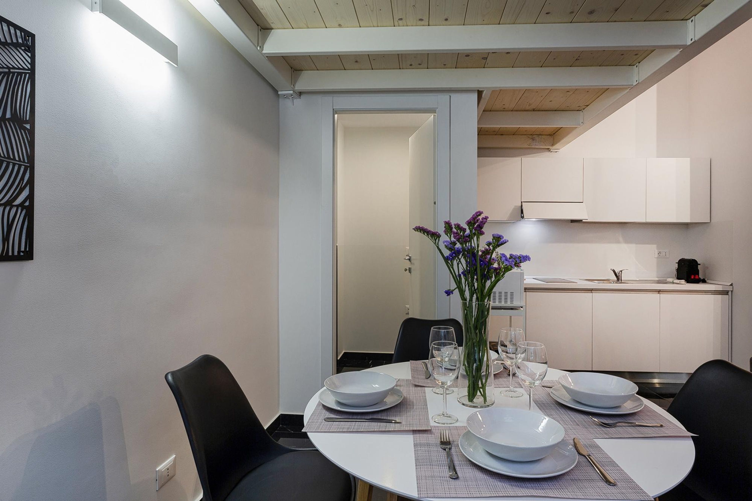 Un comedor con una mesa redonda dispuesta para cuatro, platos blancos, cubiertos y un florero con flores moradas. Se aprecia una cocina al fondo con armarios blancos y un electrodoméstico negro.