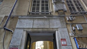 Entrada de un edificio con un arco de piedra, una placa sobre el ventanal y un cartel que dice 'AFFITTA'.