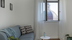 A room with a grey sofa, a small white table with a bottle and glass, and a window with a view of a dome-shaped building. - Deluxe Apartment - Avio by Wonderful Italy (Genoa)