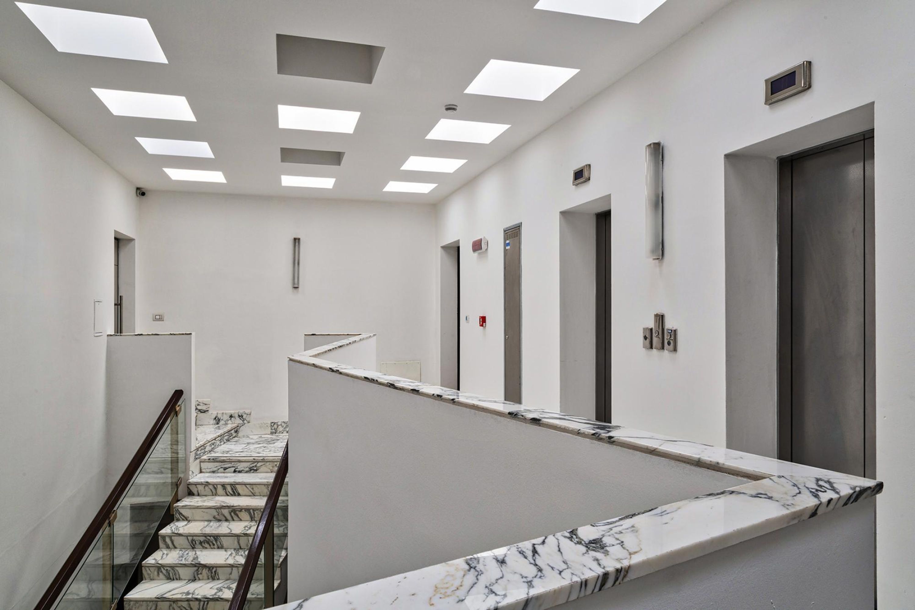 A modern hotel lobby with a marble staircase, elevators, and a glass railing.