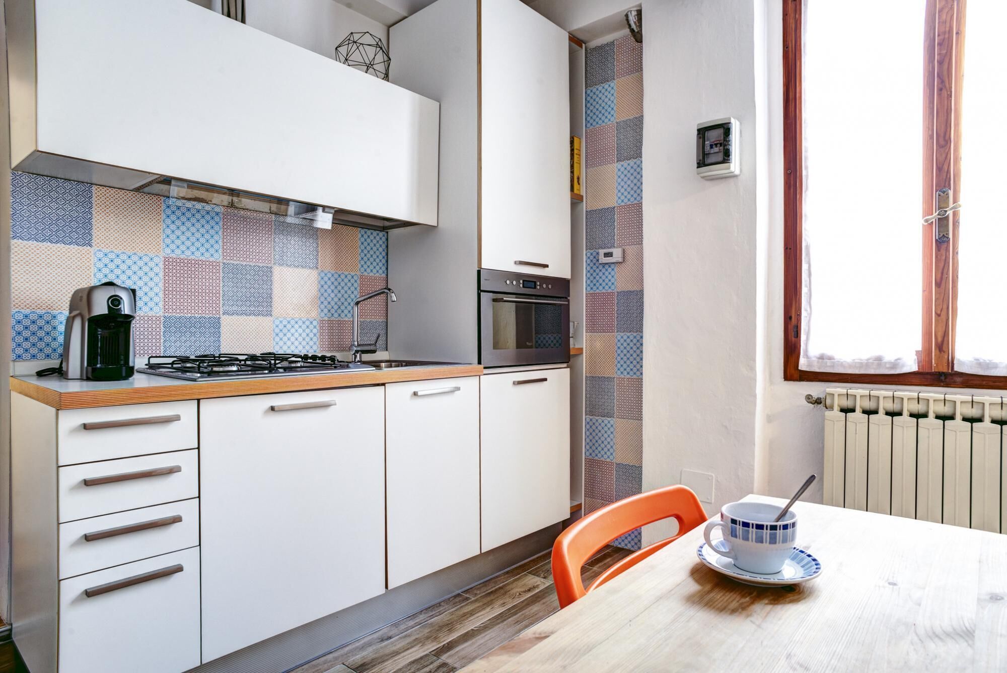 Una cocina compacta con estufa a gas, horno y cafetera. Hay una mesa de comedor con una taza y un platillo.