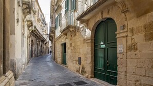 A narrow street with stone buildings, a green door, and a vent on the ground. - Duplex sul Porto Grande by Wonderful Italy (Syracuse)