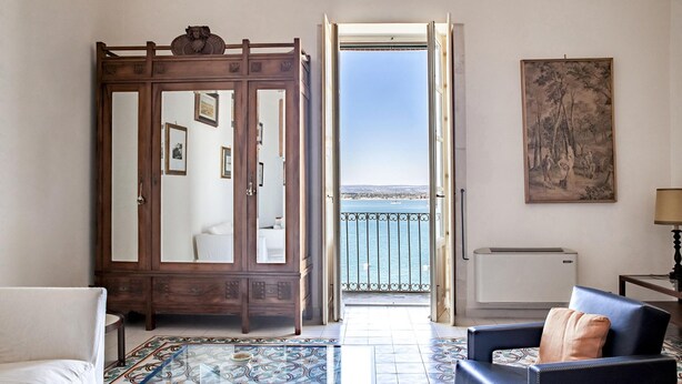 A room with a view to the sea, a wooden wardrobe, a patterned floor, and a chair with a cushion. - Duplex sul Porto Grande by Wonderful Italy (Syracuse)