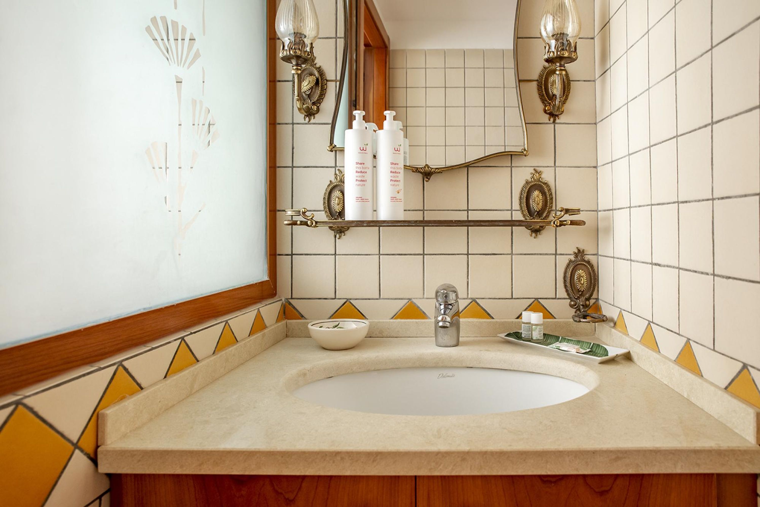 A bathroom with a white sink, wooden cabinets, and a mirror. There are toiletries on a shelf and decorative wall sconces.