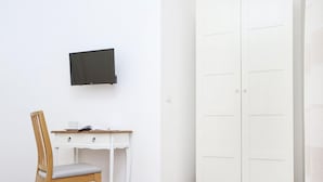 Quarto com escrivaninha branca, cadeira de madeira, televisão de tela plana na parede e guarda-roupa branco com várias portas.