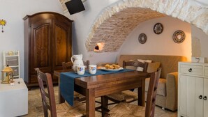 Un coin repas chaleureux avec une table en bois, des chaises et une nappe bleue. Un arc en pierre surplombe la table, un téléviseur est fixé au mur, et des assiettes décoratives ornent également le mur.