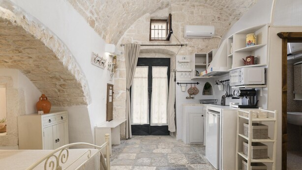 Une petite cuisine aux murs de pierre, avec des meubles blancs, une petite fenêtre et une porte donnant sur une autre pièce.