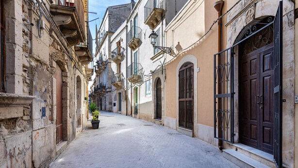 A narrow cobblestone street lined with historic buildings, featuring balconies, wrought iron gates, and a potted plant.