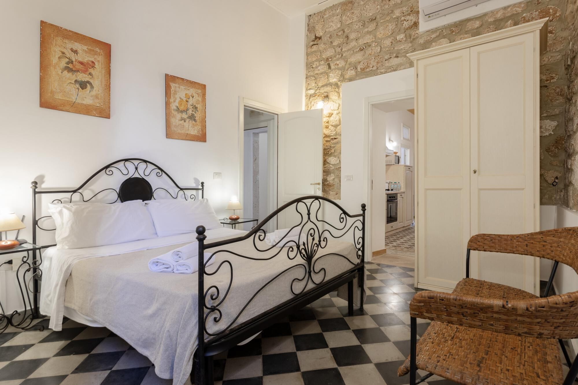 A bedroom with a checkered floor, a bed with a metal frame, two wall-mounted paintings, a wicker chair, and a stone wall.