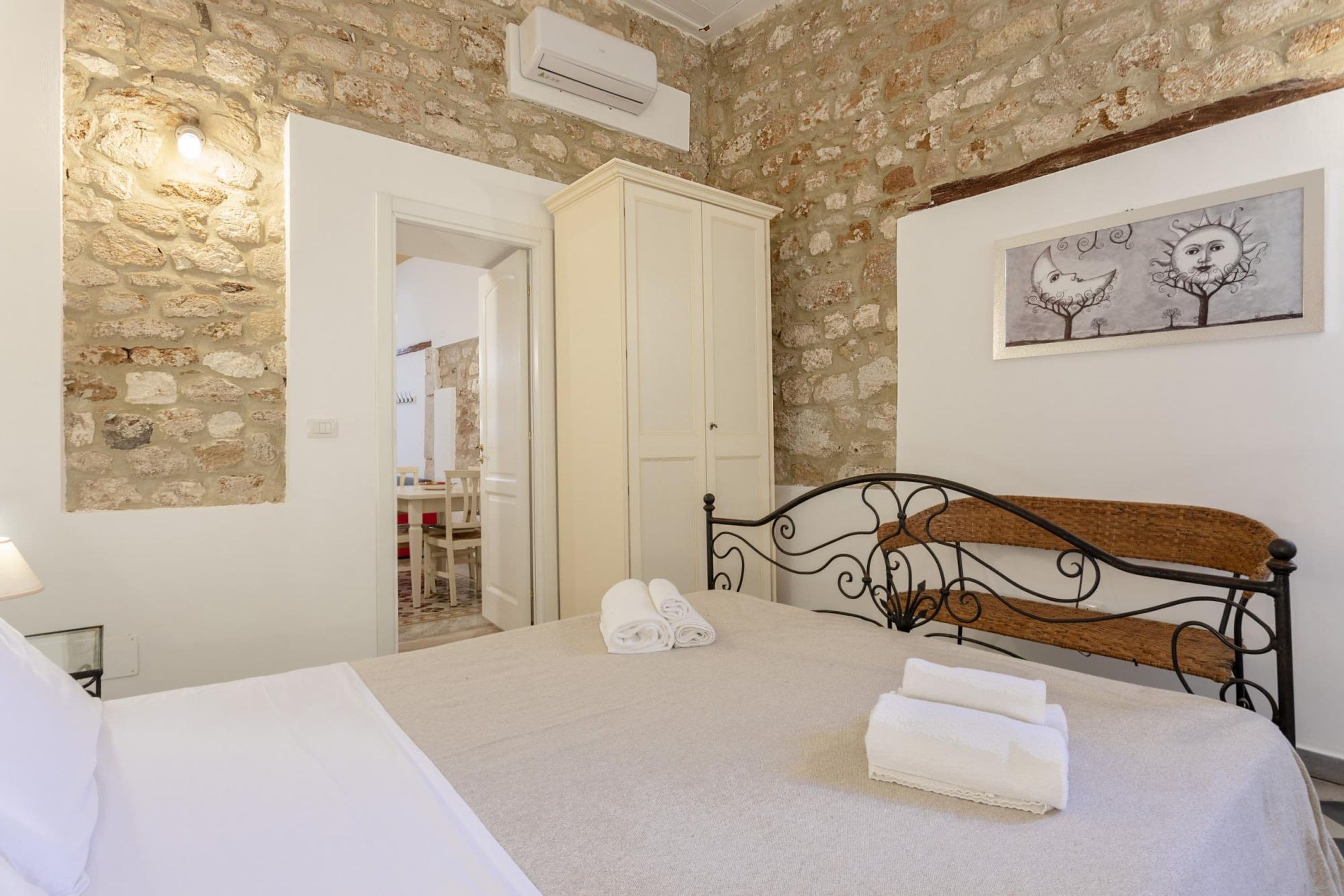 A bedroom with a stone wall, a bed with a metal headboard, a white wardrobe, and a framed artwork on the wall.