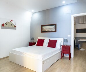 A bedroom with a white bed, red pillows, a framed picture on the wall, and a small red nightstand with a lamp.