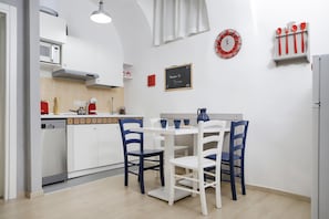 Cozinha compacta com armários brancos, micro-ondas, torradeira vermelha e chaleira azul. Há uma mesa branca com cadeiras azuis, um quadro-negro com uma mensagem de boas-vindas e um prato decorativo vermelho e branco na parede.