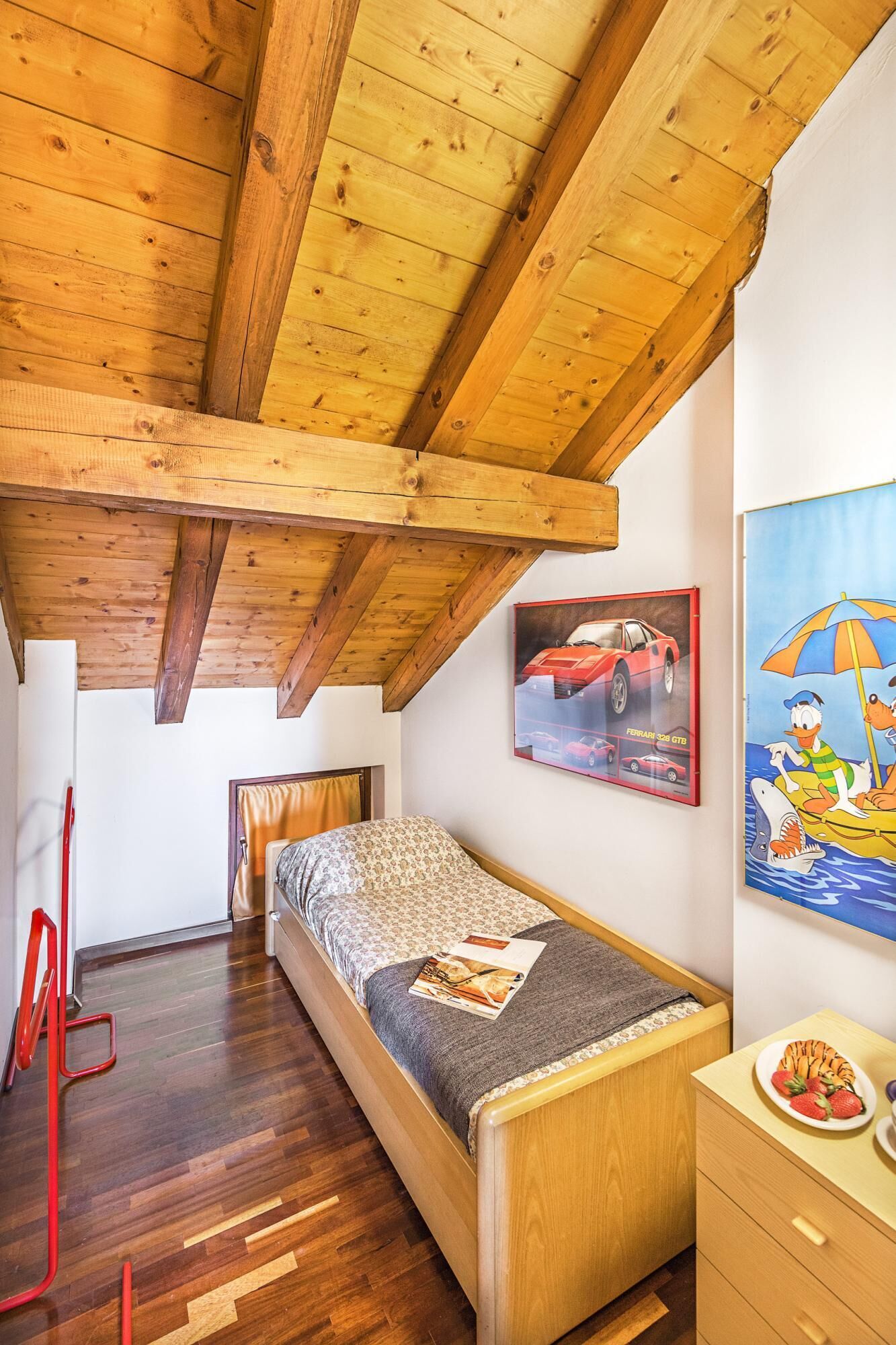A bedroom with a wooden ceiling, a bed with a patterned bedspread, a painting of a car, and a small table with a plate of strawberries.