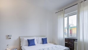 A bedroom with a bed, a nightstand, a lamp, a window with curtains, and a view of buildings outside.