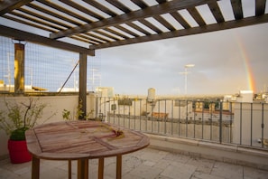 A rooftop terrace with a wooden table and chairs, a red planter, and a view of the cityscape and a rainbow.
