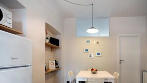 A compact kitchen with a white refrigerator, a dining table with chairs, a hanging light fixture, and a door.