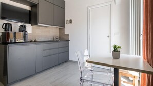 A modern kitchen with grey cabinets, a built-in oven, and a coffee machine. There is a small table with a potted plant and a clear chair.