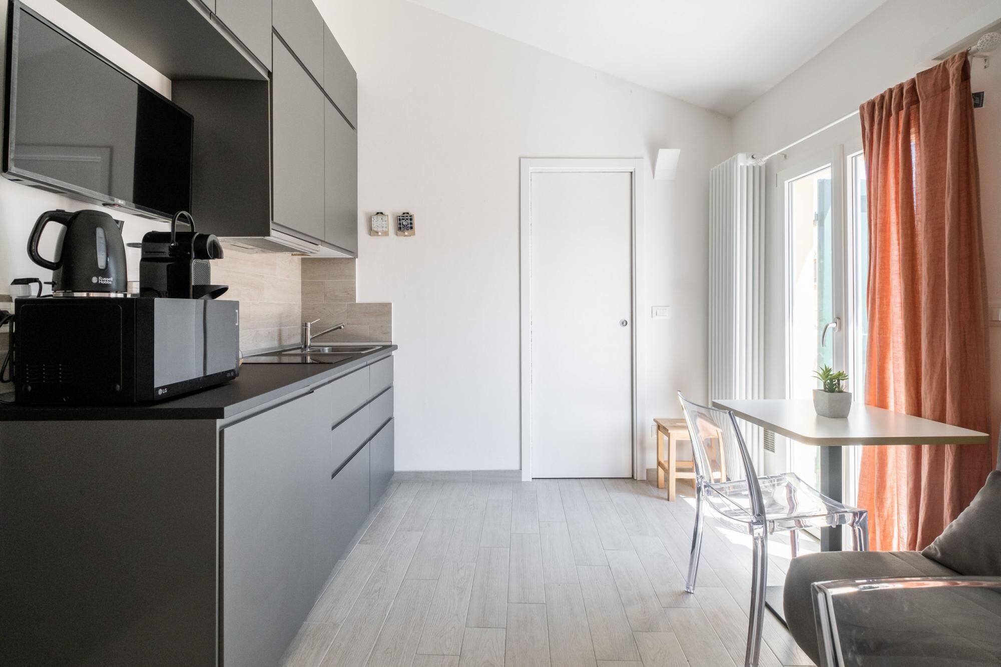 A modern kitchen with a built-in microwave, coffee maker, and kettle. There is a dining area with a table and chairs, and a window with curtains.